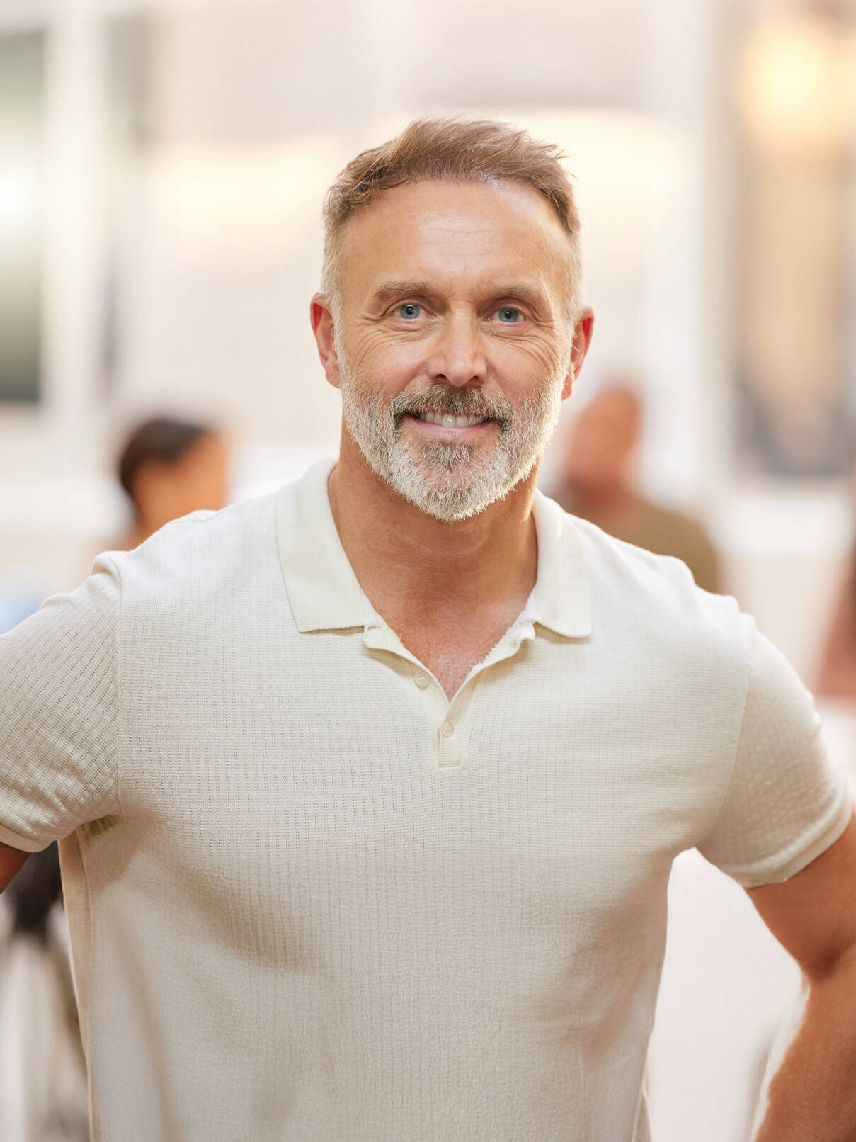 A middle-aged man with short hair and a beard, wearing a light-colored polo shirt, stands indoors with a neutral expression—showcasing the excellent results from Cincinnati hair restoration specialists in the blurred background.