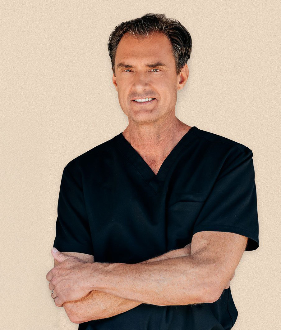 A man with short brown hair wearing black medical scrubs stands with arms crossed, smiling confidently at the camera&mdash;representing the experienced hair restoration surgeons Cincinnati trusts at Wolf Hair Restoration Cincinnati.