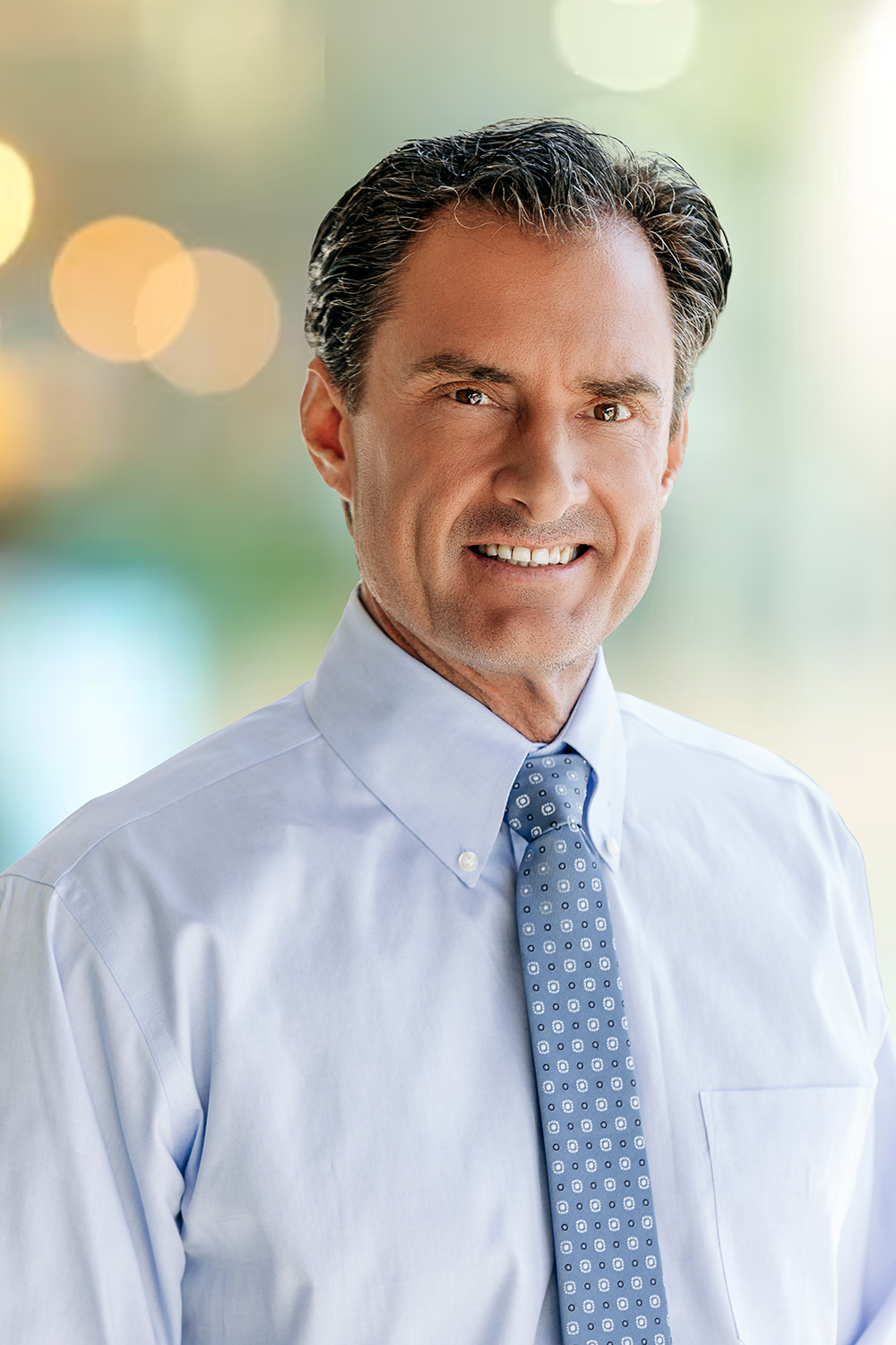 A middle-aged man with dark hair, thanks to Wolf Hair Restoration Cincinnati, wearing a light blue dress shirt and patterned tie, smiling at the camera with a blurred background.