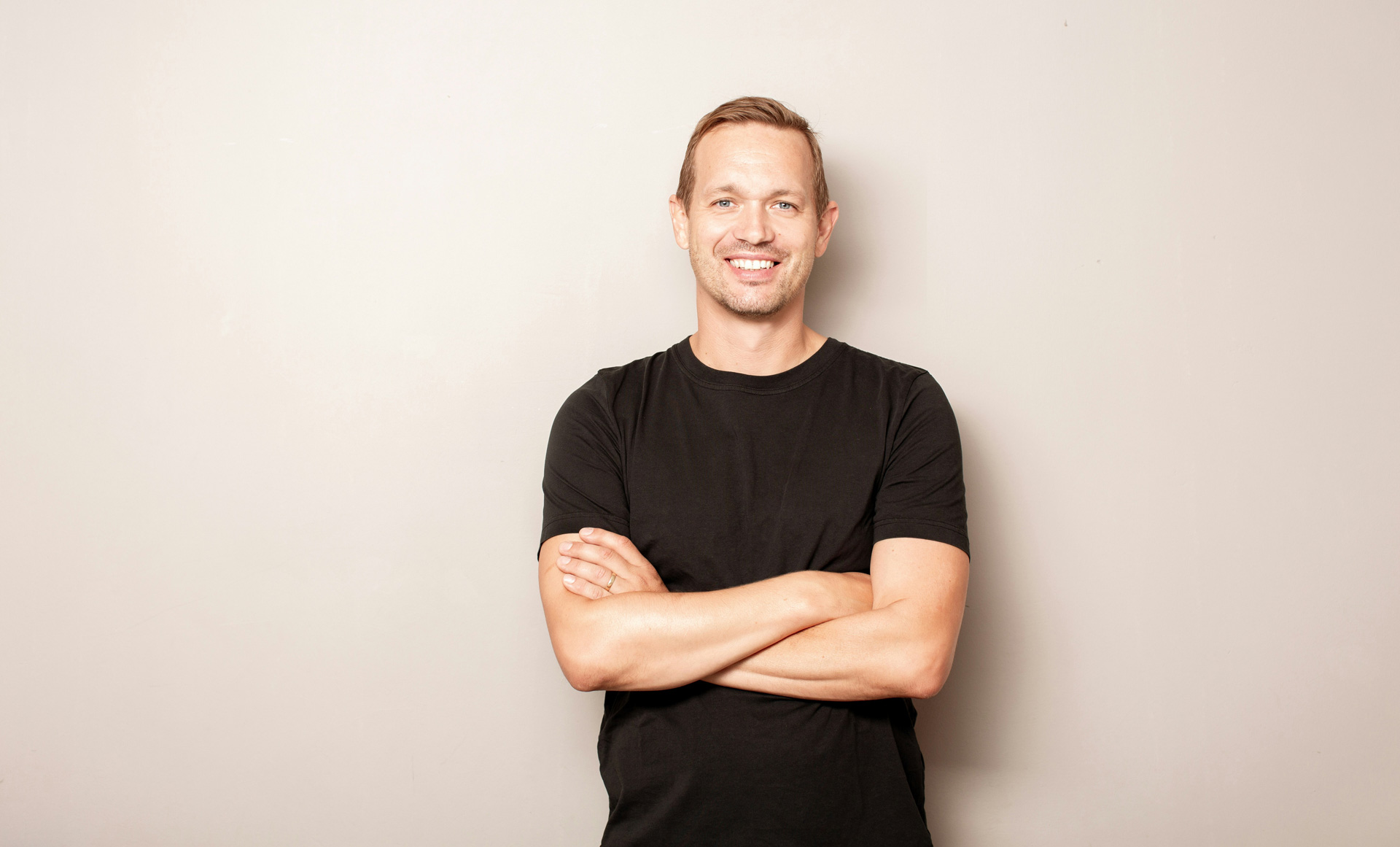 A man in a black t-shirt stands against a plain light-colored wall with his arms crossed, smiling at the camera.