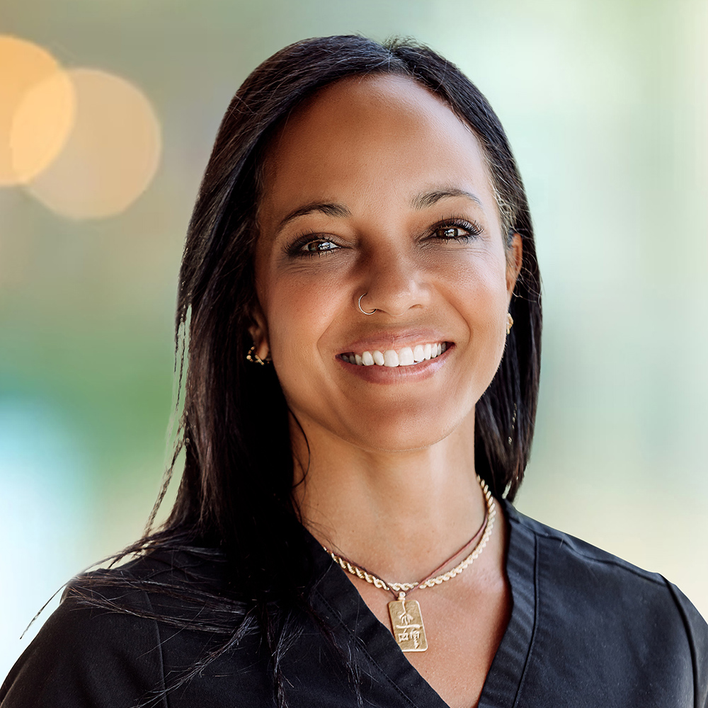 A woman with long dark hair&mdash;thanks to Wolf Hair Restoration Cincinnati&mdash;wears a black top and gold jewelry, smiling at the camera against a blurred outdoor background.