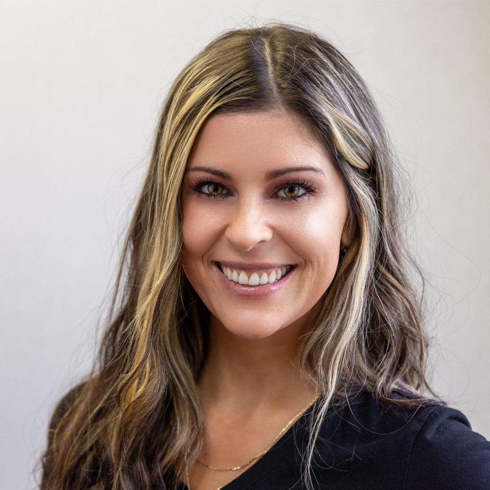 Woman with long brown hair and blonde highlights, styled by experienced hair restoration surgeons at Wolf Hair Restoration Cincinnati, wearing a black top and gold necklace, smiling at the camera against a plain light background.