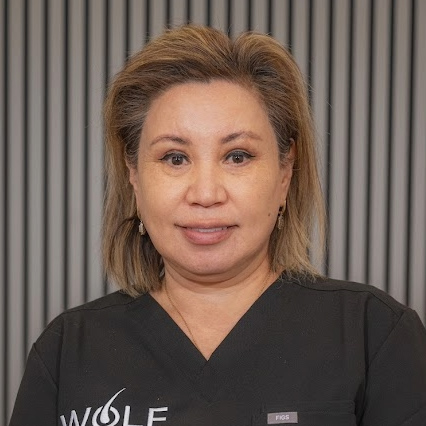 A woman with light brown hair wearing black scrubs stands in front of a gray vertical striped background, facing the camera and smiling slightly&mdash;representing the caring team at Wolf Hair Restoration Cincinnati.