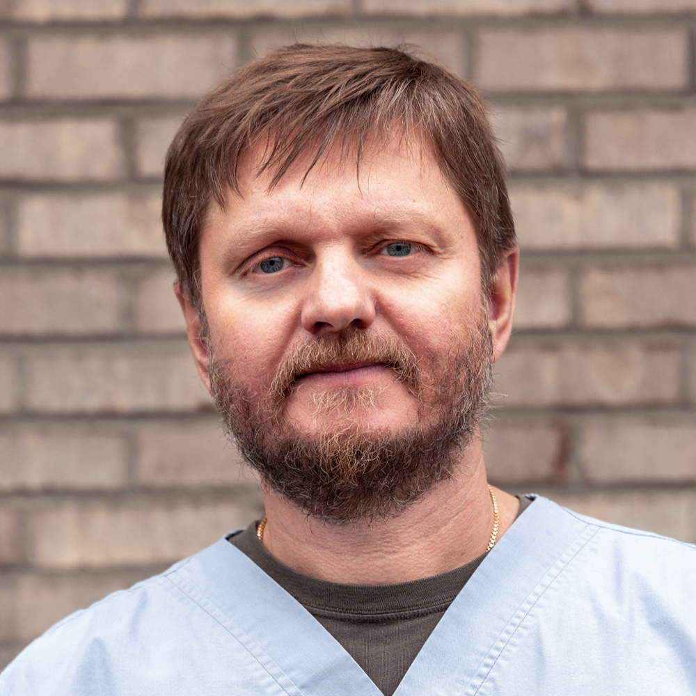 A man with short brown hair and a beard, wearing light blue scrubs, stands in front of a brick wall, representing the experienced hair restoration surgeons at Wolf Hair Restoration Cincinnati.