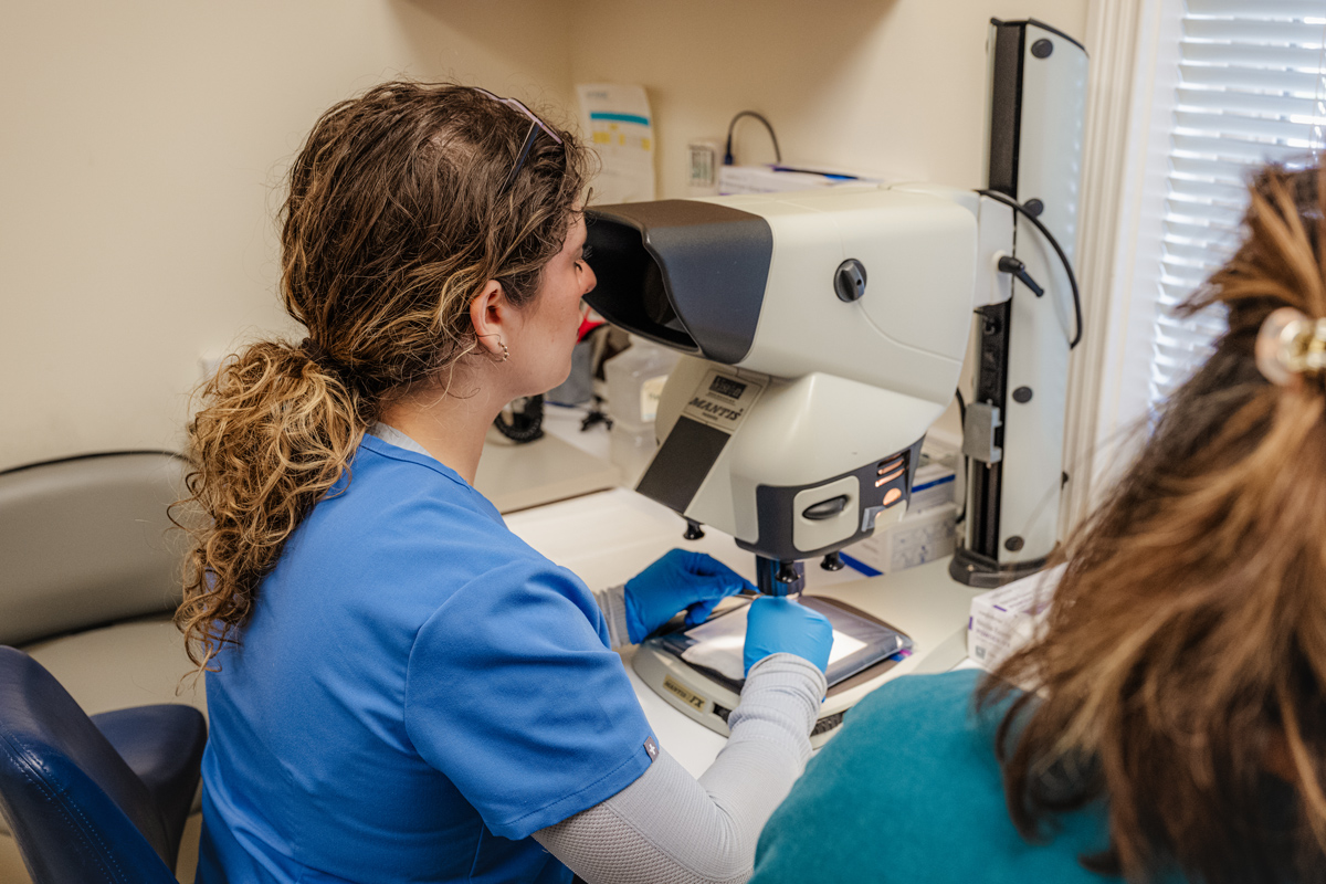A person wearing blue scrubs and gloves examines a sample using a microscope in a medical or laboratory setting, similar to procedures used in Women&rsquo;s Hair Restoration Cincinnati clinics.