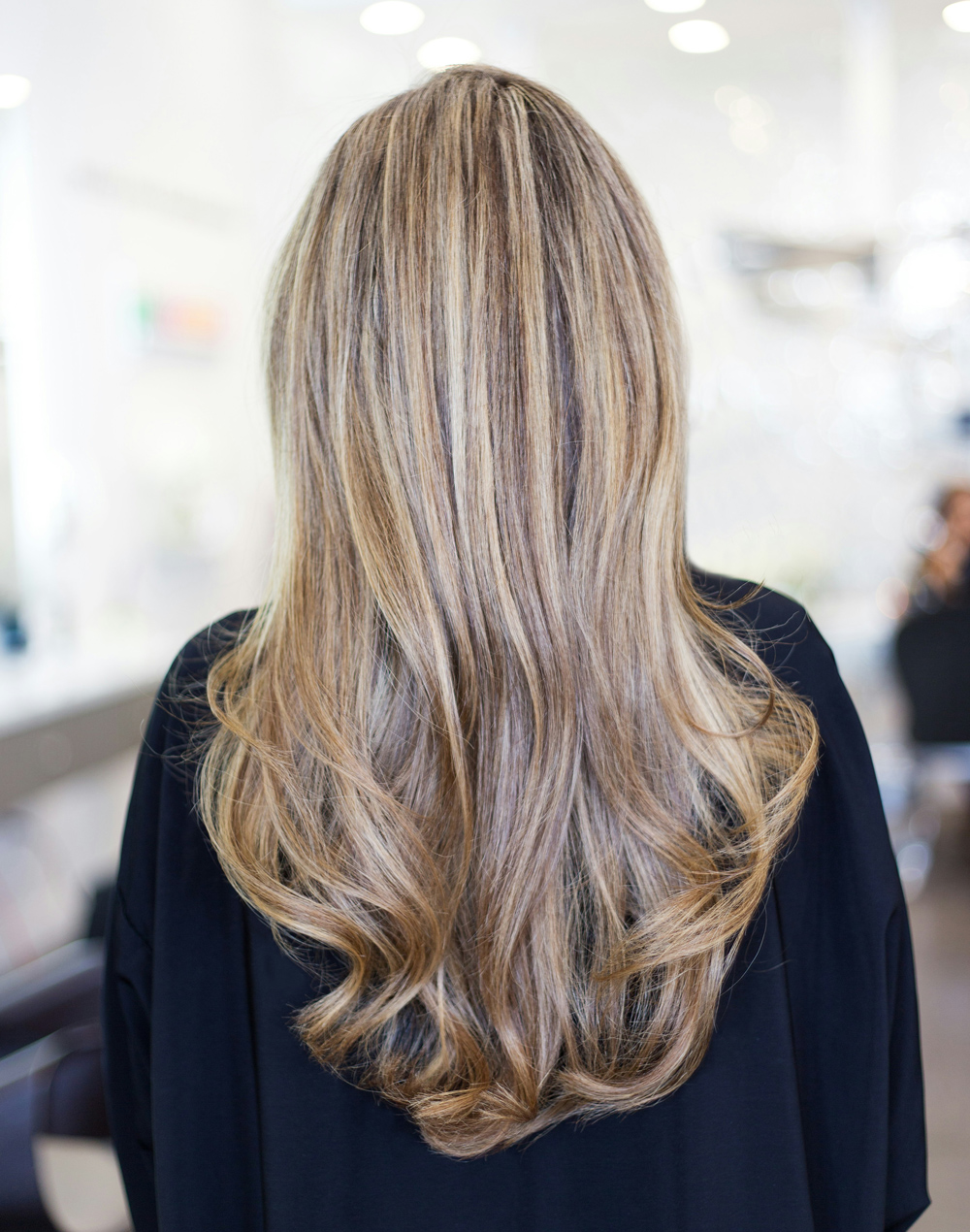 A person with long, wavy, blonde hair with highlights is shown from the back in a brightly lit salon, showcasing the results of Women&rsquo;s Hair Restoration Cincinnati.
