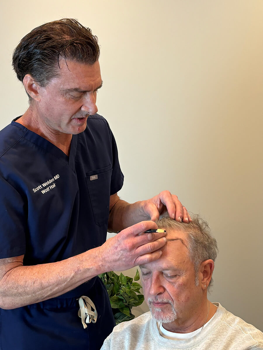 A medical professional in navy scrubs examines the scalp of a seated older man using a handheld device, providing personalized care as part of the Wolf Hair Restoration FAQ process.