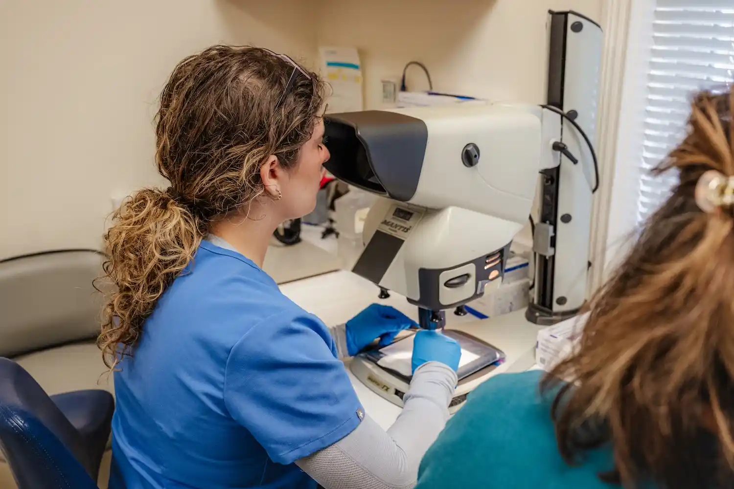 A healthcare professional in blue scrubs uses a medical imaging device while another person is partially visible beside her.