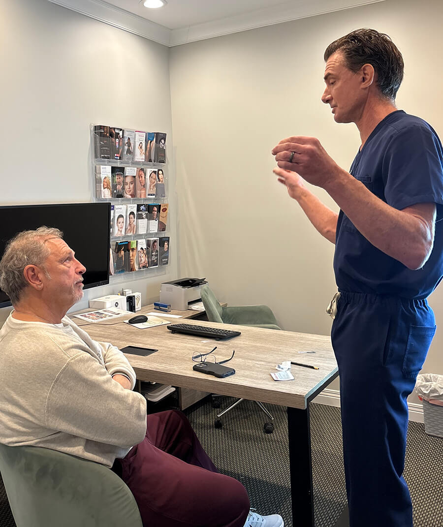 A doctor in blue scrubs stands and explains something to a seated older man in a medical office setting, addressing his questions from the Wolf Hair Restoration FAQ.