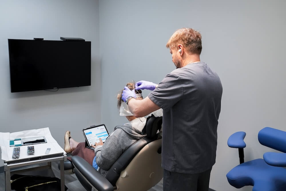 A medical professional examines a seated patient&rsquo;s scalp in a clinic room; the patient uses a tablet while the provider wears gloves.