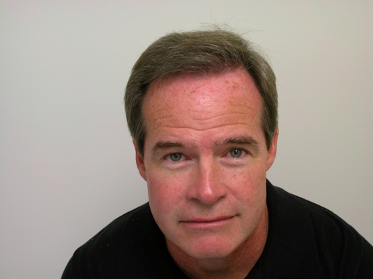 A middle-aged man with short brown hair and a neutral expression looks up at the camera against a plain light-colored background.